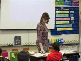 teacher in front of whiteboard instructing class and pointing to a book on a students desk
